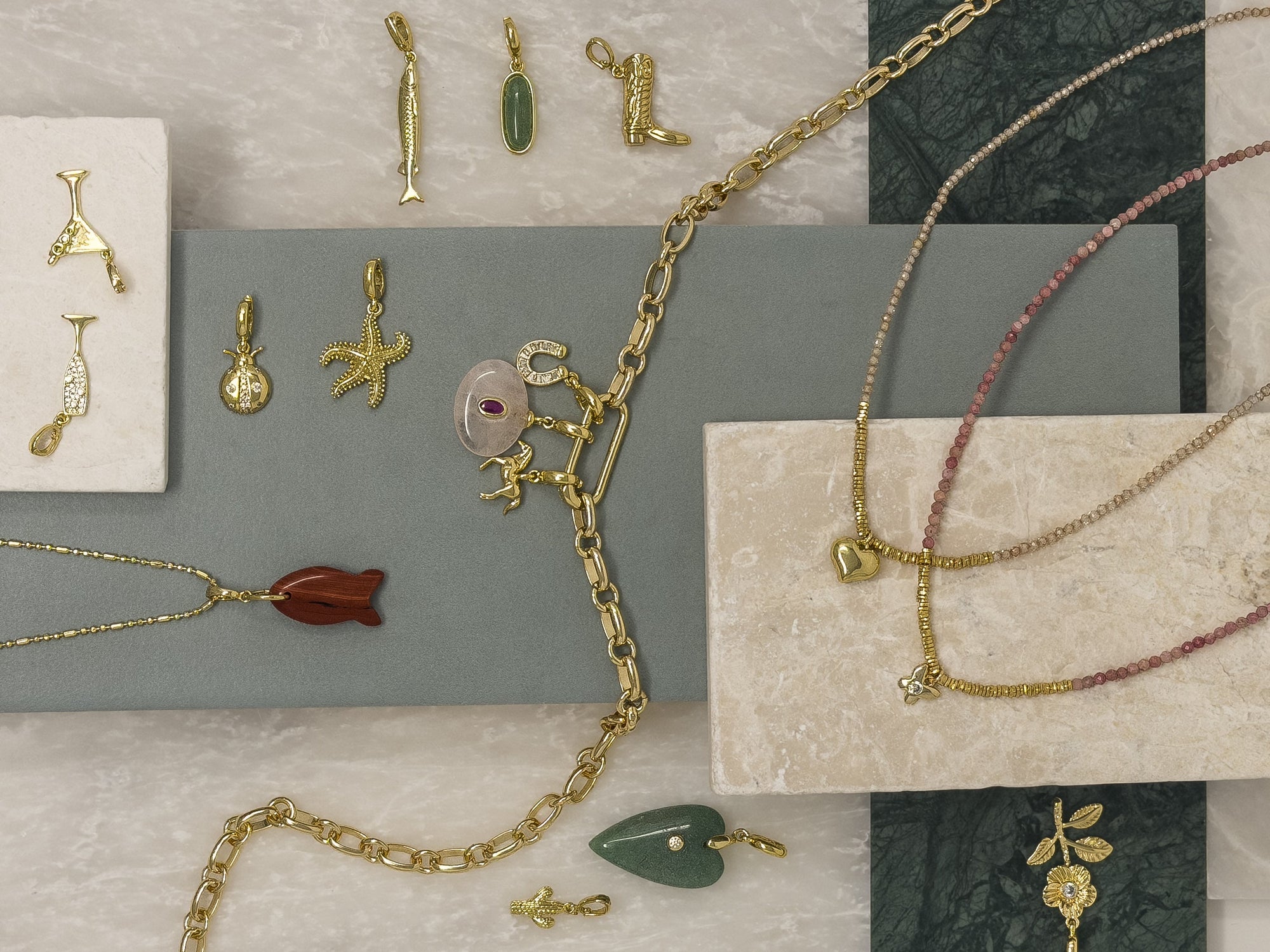 A styled flat-lay of multiple gold-tone jewelry pieces, including necklaces, bracelets, and earrings, arranged on neutral stone and slate blocks.