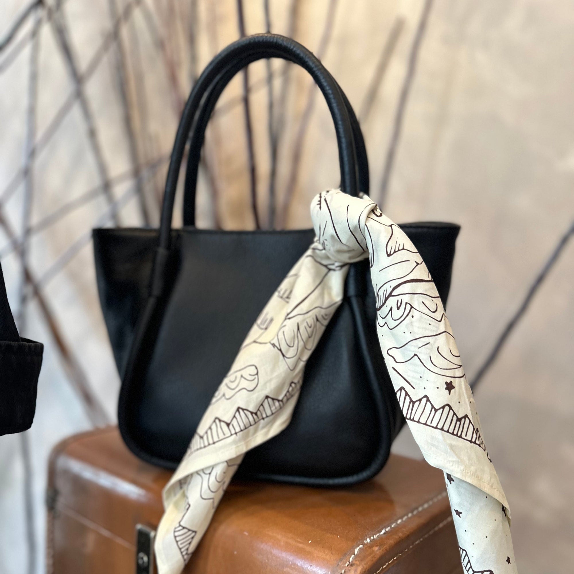 Black handbag with a white patterned scarf draped over it, placed on a brown suitcase.